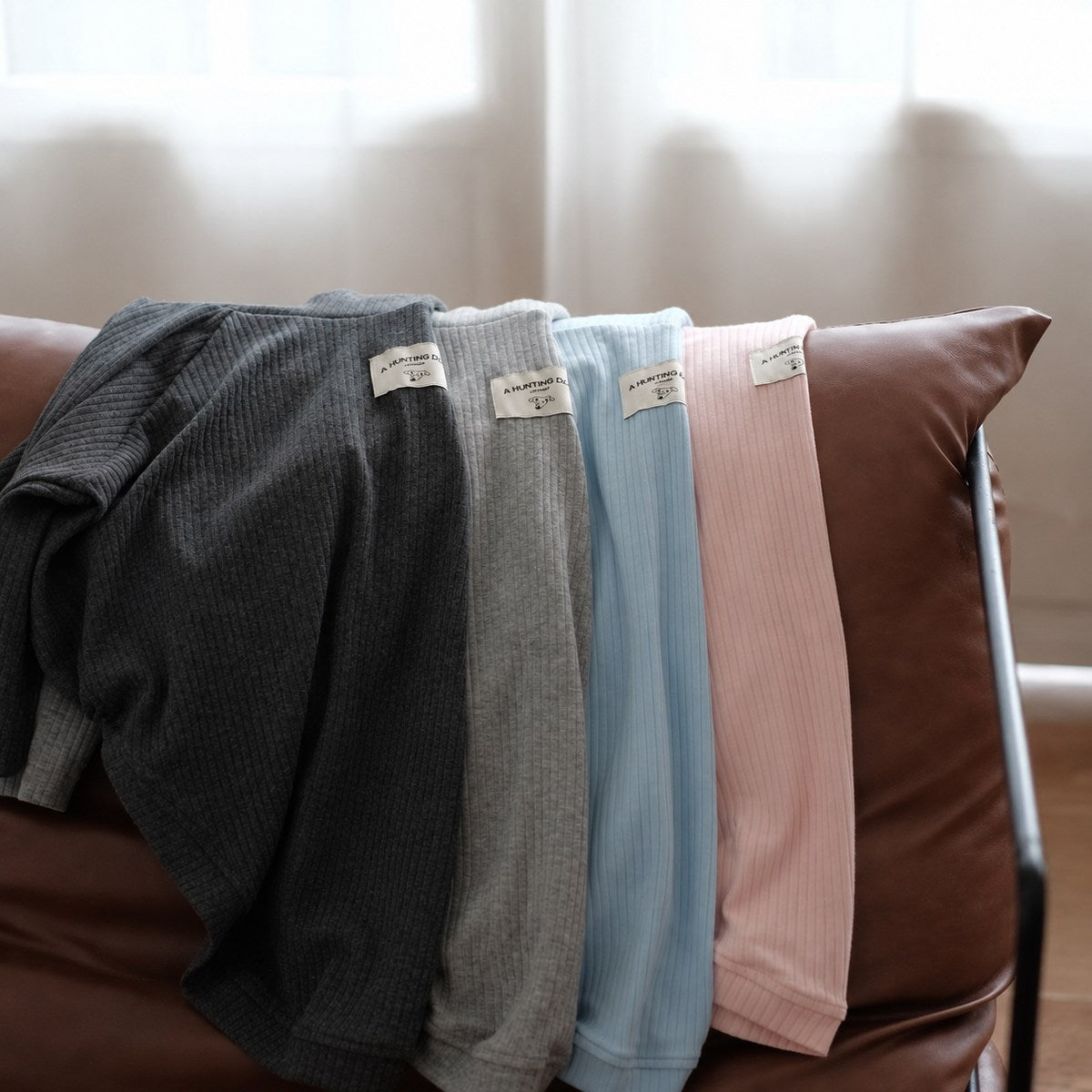 Five muted sighthound turtleneck colours arranged together over a leather chair beside a window.
